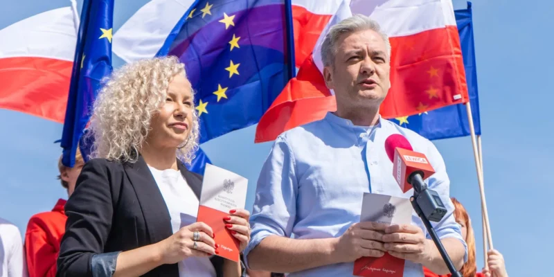 Dwóch mówców na wydarzeniu politycznym w Polsce: mężczyzna w jasnoniebieskiej koszuli przemawia do mikrofonu, trzymając kartkę z godłem, a obok niego stoi kobieta z broszurą; w tle widoczne flagi Polski i Unii Europejskiej.