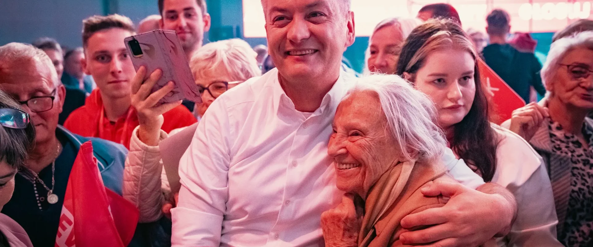 Uśmiechnięty mężczyzna w białej koszuli obejmuje starszą kobietę, otoczony przez tłum ludzi z flagami podczas publicznego spotkania.