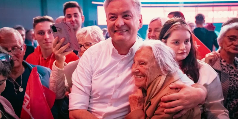 Uśmiechnięty mężczyzna w białej koszuli obejmuje starszą kobietę, otoczony przez tłum ludzi z flagami podczas publicznego spotkania.