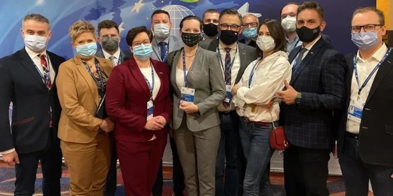 Grupa osób w eleganckich strojach, noszących maski ochronne, pozująca wspólnie na tle kolorowego tła podczas wydarzenia.