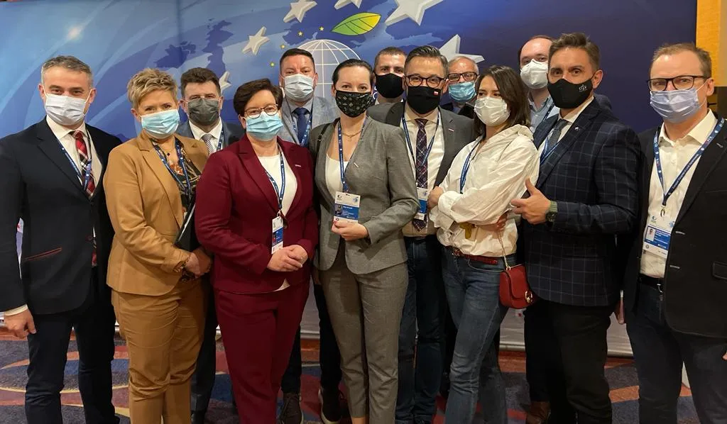 Grupa osób w eleganckich strojach, noszących maski ochronne, pozująca wspólnie na tle kolorowego tła podczas wydarzenia.