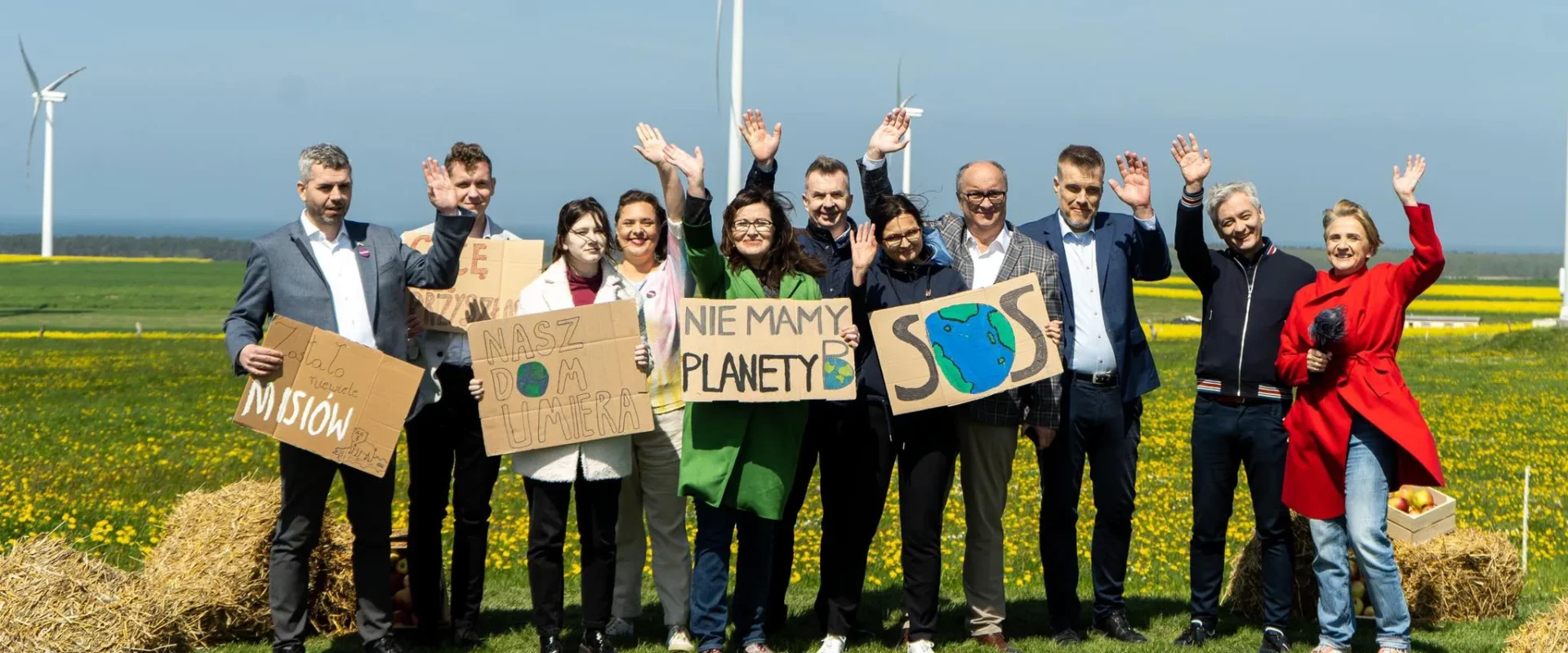 Grupa ludzi na tle wiatraków i zielonej łąki trzyma transparenty z hasłami ekologicznymi, wyrażając troskę o planetę.
