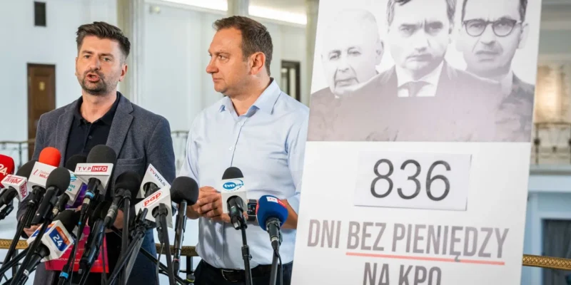 Dwóch mężczyzn przemawia na konferencji prasowej otoczonych mikrofonami, a w tle widnieje plakat z napisem „836 dni bez pieniędzy na KPO”.