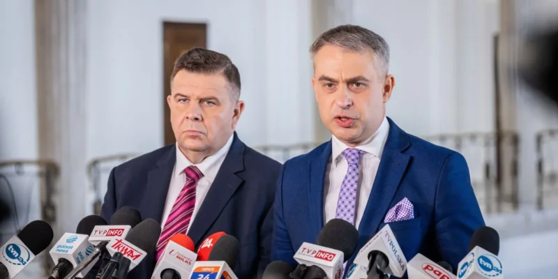 Dwóch mężczyzn w garniturach podczas konferencji prasowej, stojących za licznymi mikrofonami z logotypami mediów, w eleganckim wnętrzu z kolumnami.