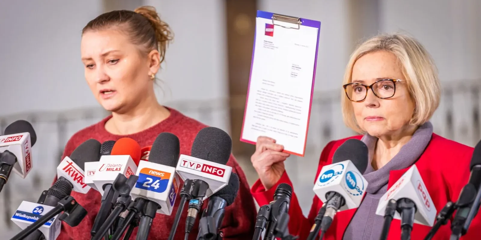 Na konferencji prasowej dwie kobiety stoją wśród licznych mikrofonów; jedna z nich trzyma kartkę z dokumentem.