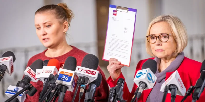 Na konferencji prasowej dwie kobiety stoją wśród licznych mikrofonów; jedna z nich trzyma kartkę z dokumentem.