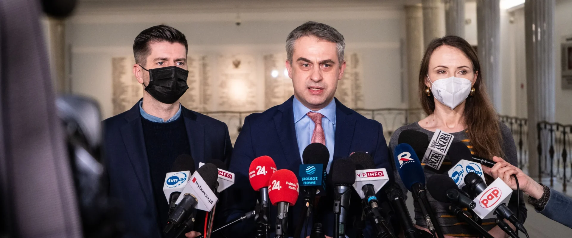 Na zdjęciu trzech polityków stoi przed mikrofonami w tle, w trakcie konferencji prasowej.