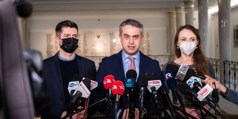 Na zdjęciu trzech polityków stoi przed mikrofonami w tle, w trakcie konferencji prasowej.