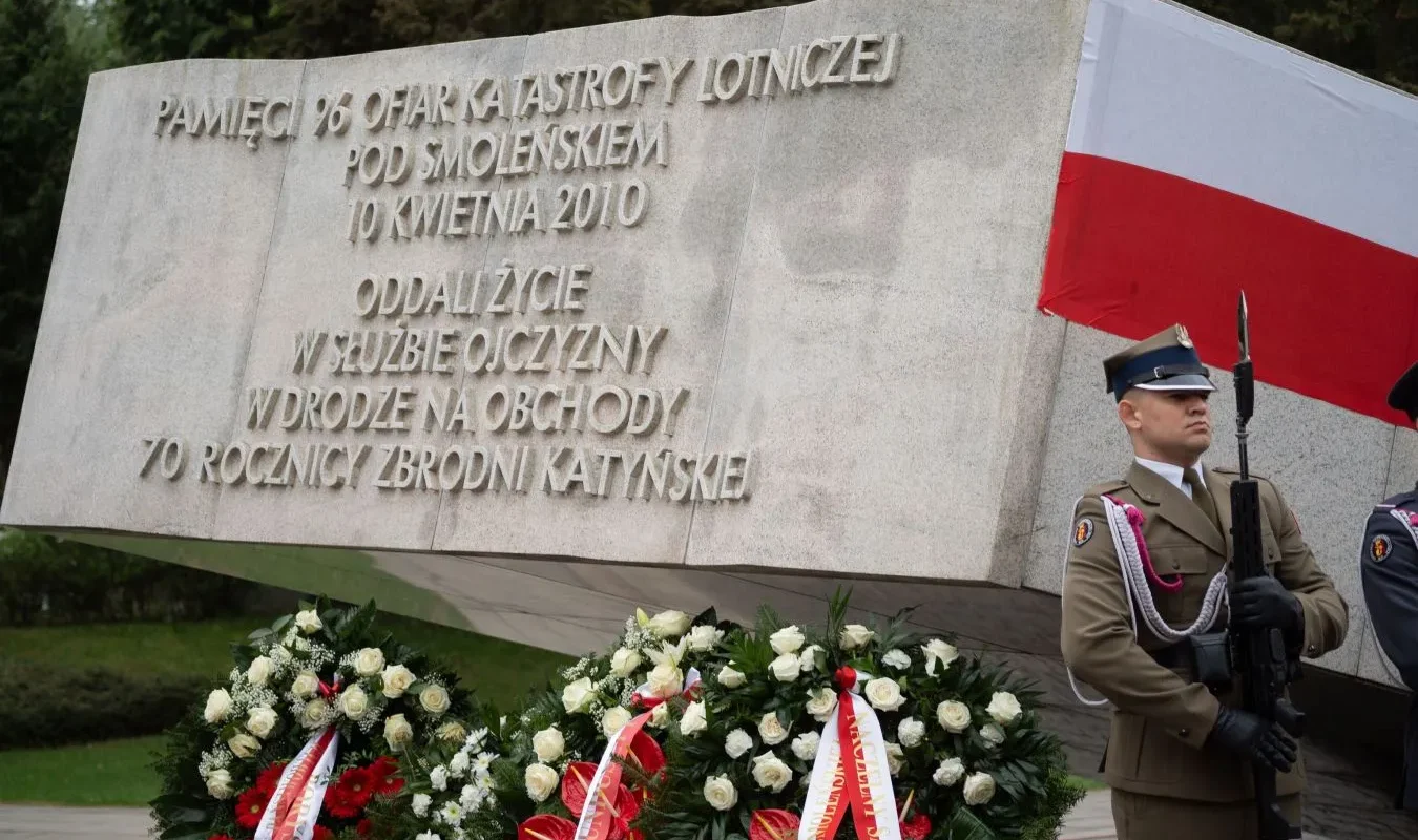 Kamienny pomnik upamiętniający ofiary katastrofy lotniczej pod Smoleńskiem z 2010 roku, otoczony wieńcami i stojący obok żołnierzy w mundurach z biało-czerwoną flagą w tle.