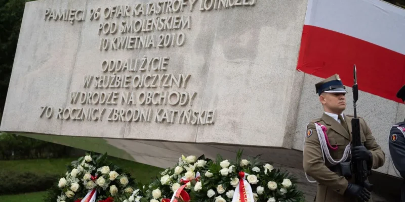 Kamienny pomnik upamiętniający ofiary katastrofy lotniczej pod Smoleńskiem z 2010 roku, otoczony wieńcami i stojący obok żołnierzy w mundurach z biało-czerwoną flagą w tle.