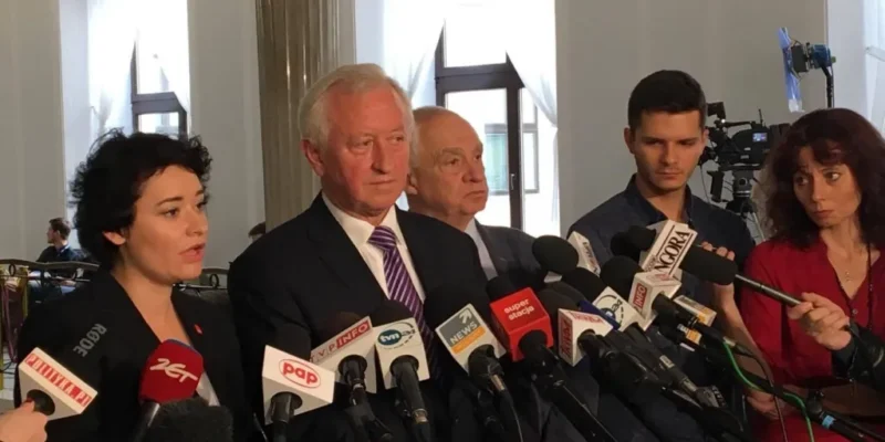 Kilka osób podczas konferencji prasowej stoi przy stojakach z licznymi mikrofonami, a dziennikarze z kamerami nagrywają ich wypowiedzi w jasnym wnętrzu.