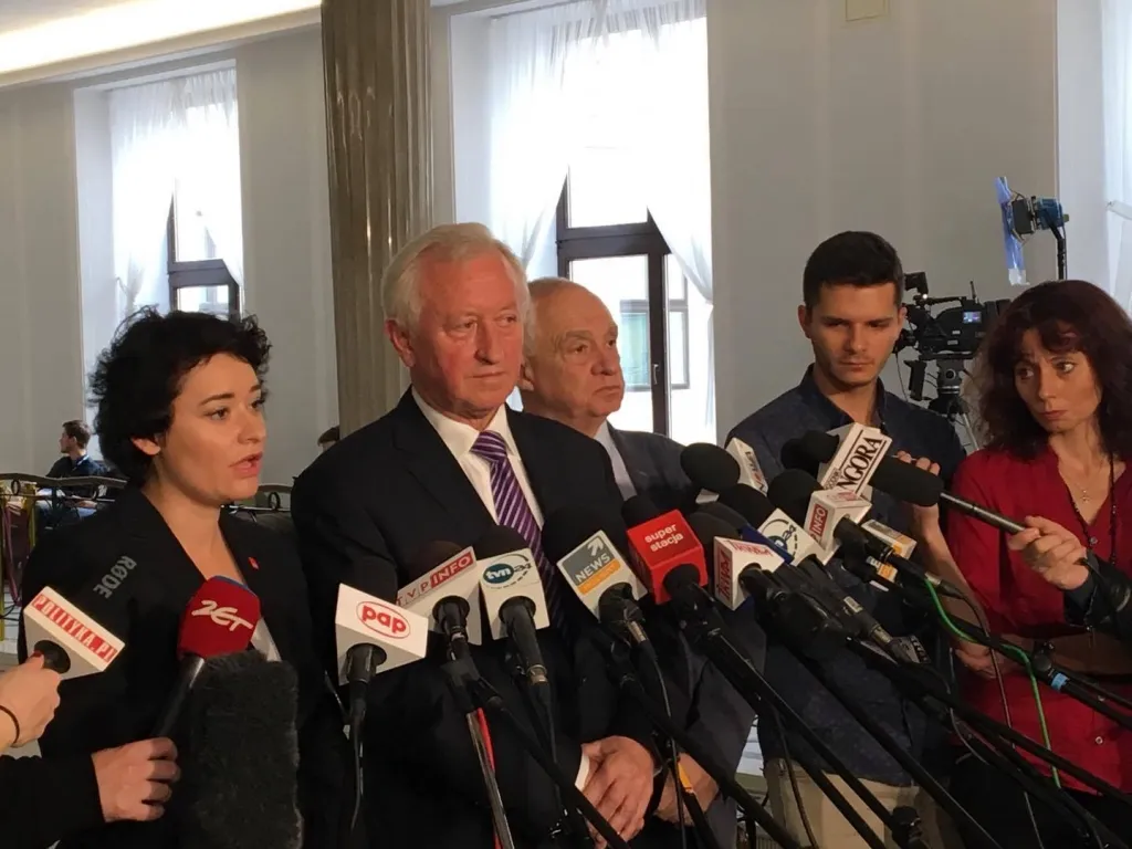 Kilka osób podczas konferencji prasowej stoi przy stojakach z licznymi mikrofonami, a dziennikarze z kamerami nagrywają ich wypowiedzi w jasnym wnętrzu.