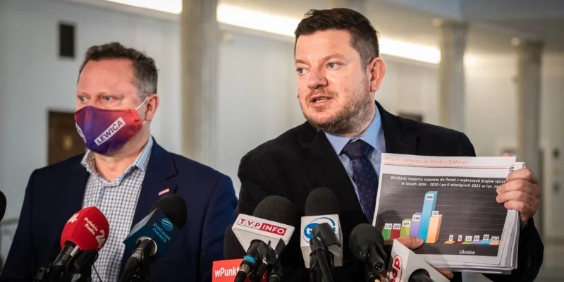 Dwóch mężczyzn w garniturach na konferencji prasowej, z których jeden trzyma plik z wykresami dotyczącymi importu cementu z Białorusi, a drugi nosi maseczkę z napisem LEWICA, przed nimi stoją liczne mikrofony.