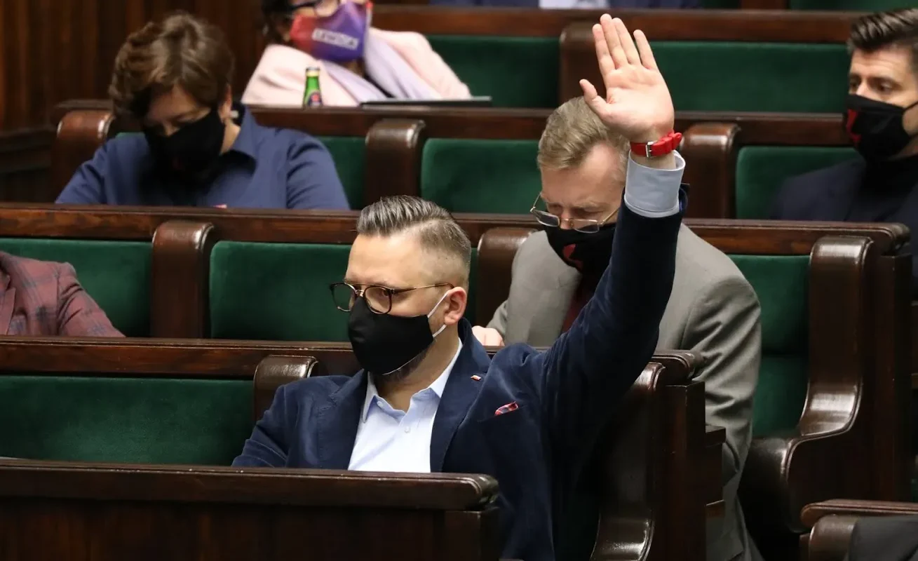 Mężczyzna w granatowym garniturze i maseczce podnosi rękę podczas posiedzenia Sejmu, siedząc wśród innych uczestników też w maseczkach.
