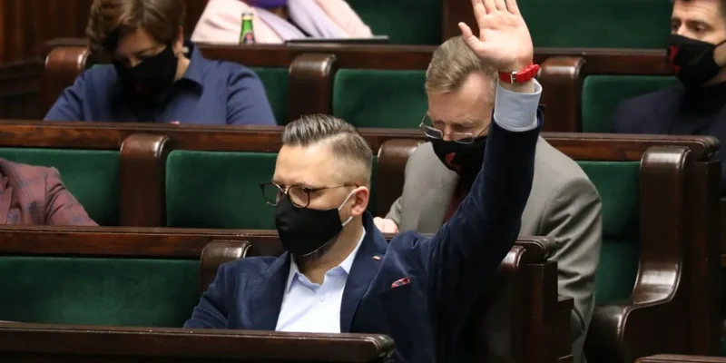 Mężczyzna w granatowym garniturze i maseczce podnosi rękę podczas posiedzenia Sejmu, siedząc wśród innych uczestników też w maseczkach.