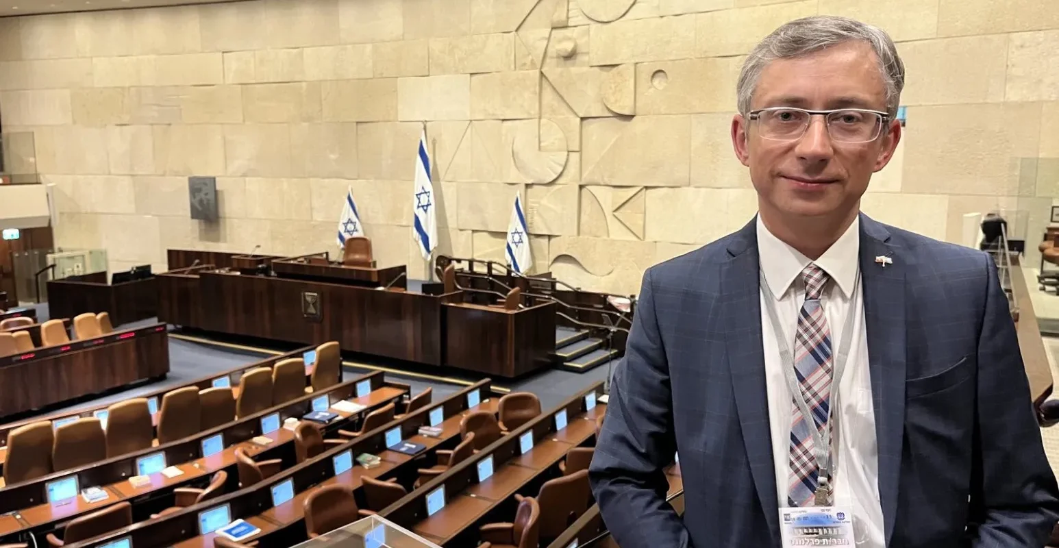 Na zdjęciu widać mężczyznę w garniturze stojącego w sali obrad z widokiem na ławki i flagi Izraela.