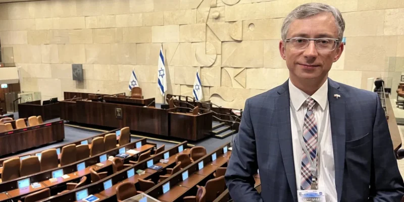 Na zdjęciu widać mężczyznę w garniturze stojącego w sali obrad z widokiem na ławki i flagi Izraela.