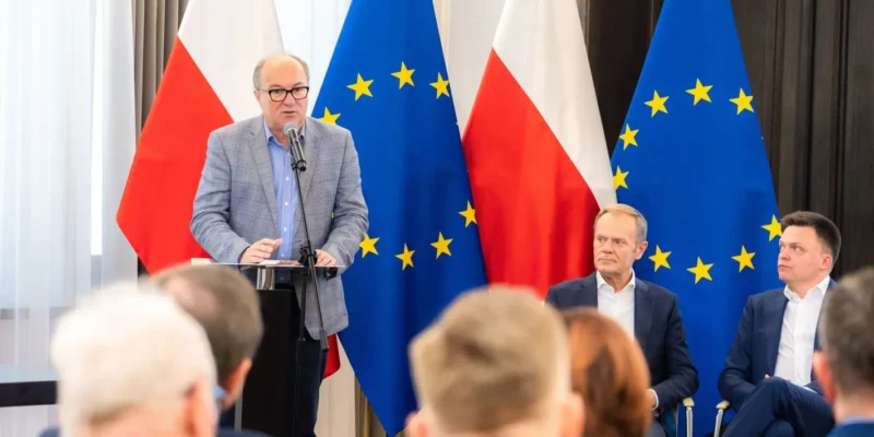 Mężczyzna w garniturze przemawia z podium podczas konferencji Obywatelska Kontrola Wyborów, na tle flag Polski i Unii Europejskiej oraz dwóch siedzących uczestników.