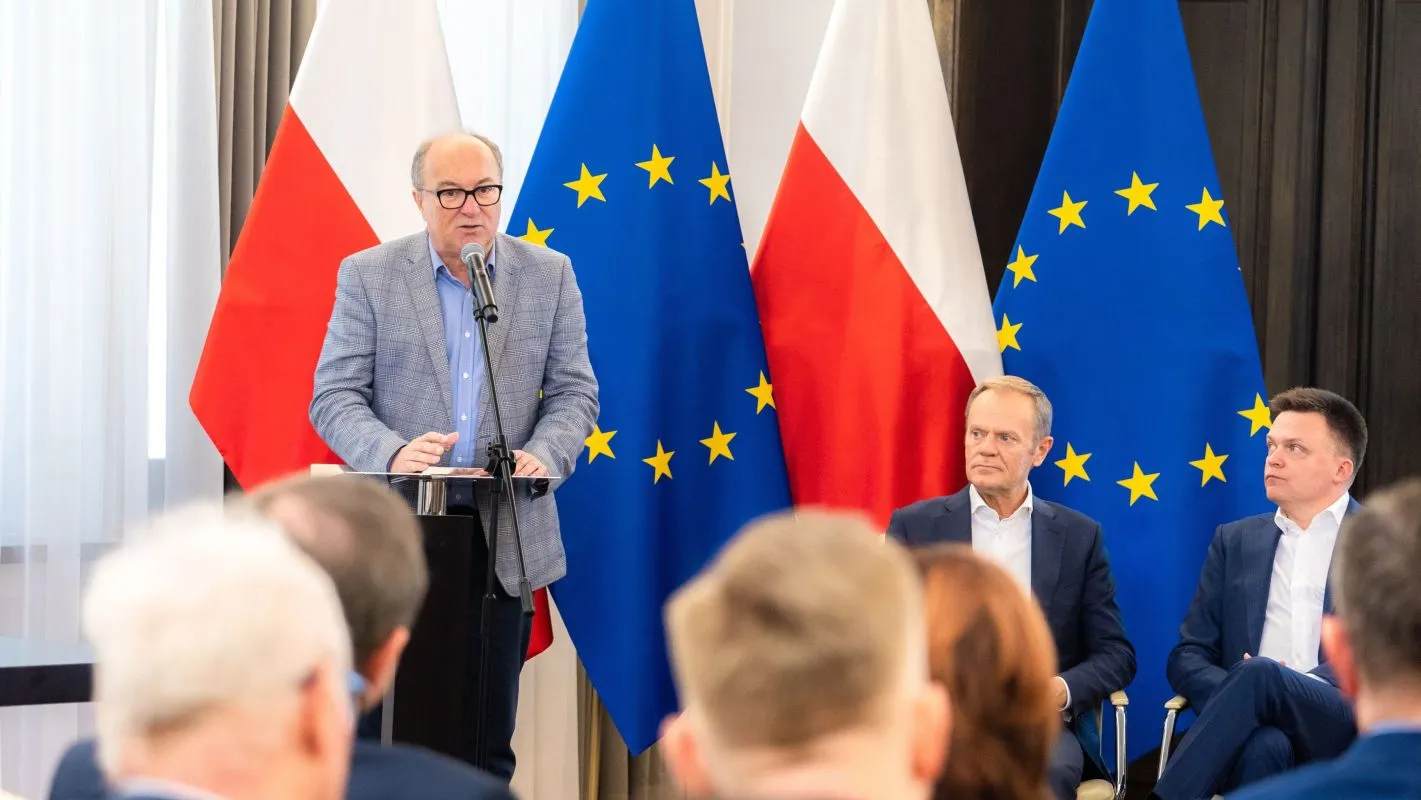 Mężczyzna w garniturze przemawia z podium podczas konferencji Obywatelska Kontrola Wyborów, na tle flag Polski i Unii Europejskiej oraz dwóch siedzących uczestników.
