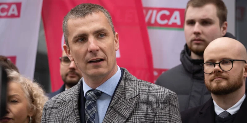 Mężczyzna w kraciastym garniturze przemawia do tłumu podczas wydarzenia politycznego, z czerwonymi flagami w tle.