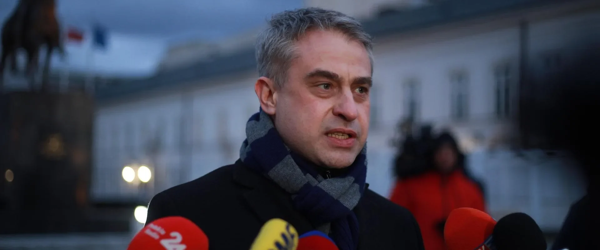 Mężczyzna w czarnym płaszczu i szaliku przemawia do mediów podczas konferencji prasowej, stojąc obok licznych mikrofonów, z tyłu widać rzeźbę konia i budynek.