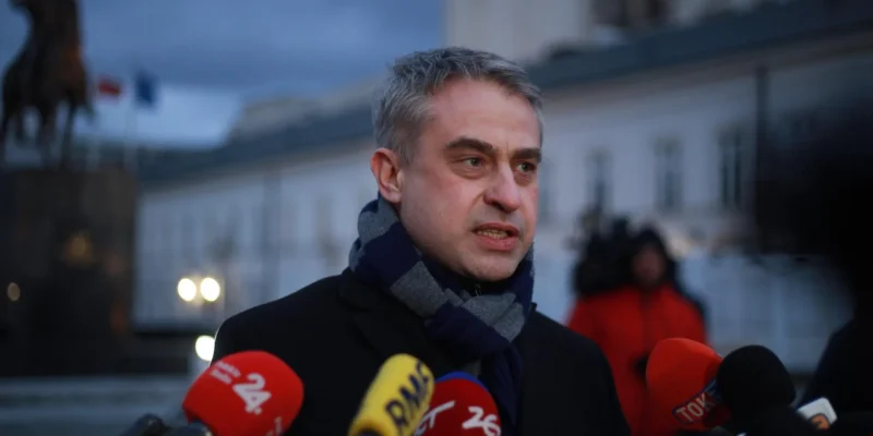 Mężczyzna w czarnym płaszczu i szaliku przemawia do mediów podczas konferencji prasowej, stojąc obok licznych mikrofonów, z tyłu widać rzeźbę konia i budynek.