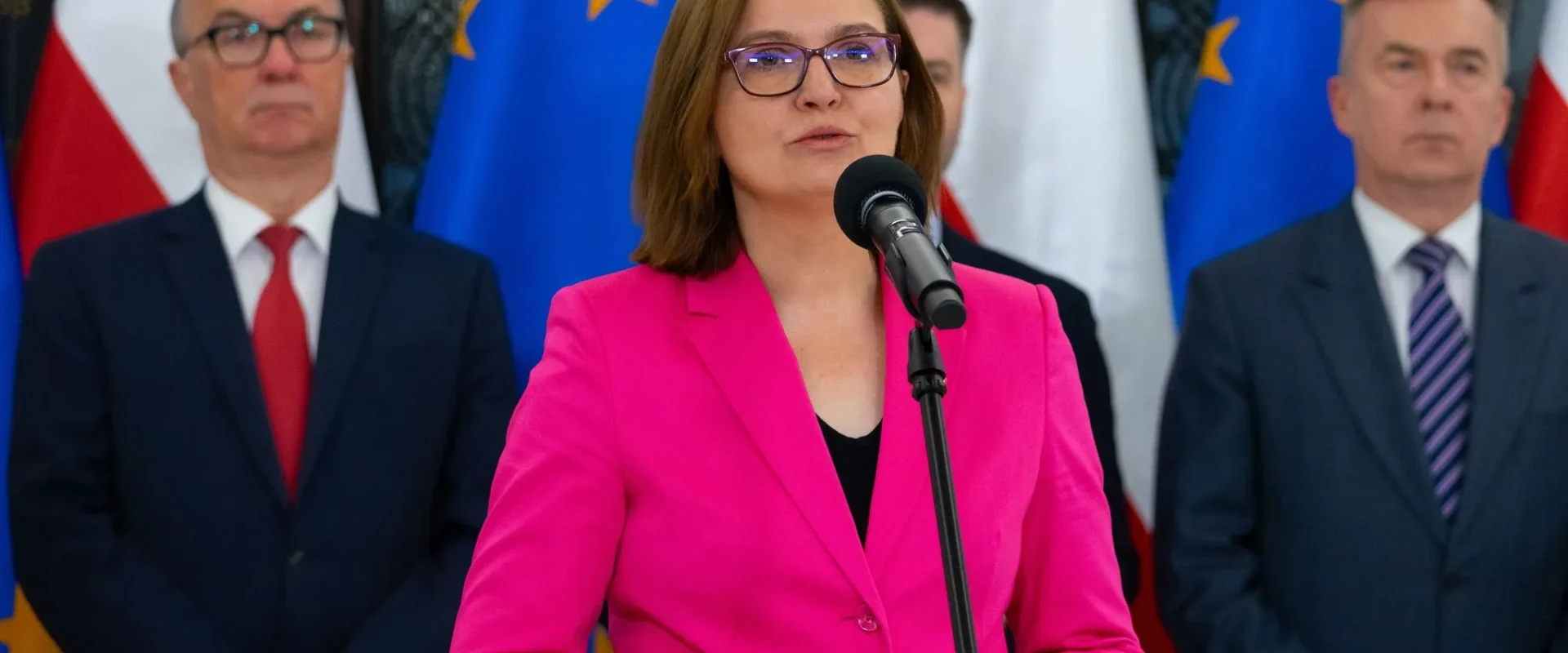 Kobieta w różowym żakiecie przemawia do mikrofonu podczas konferencji prasowej, w tle stoją urzędnicy i flagi Unii Europejskiej oraz Polski.