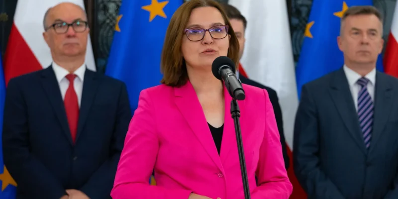 Kobieta w różowym żakiecie przemawia do mikrofonu podczas konferencji prasowej, w tle stoją urzędnicy i flagi Unii Europejskiej oraz Polski.