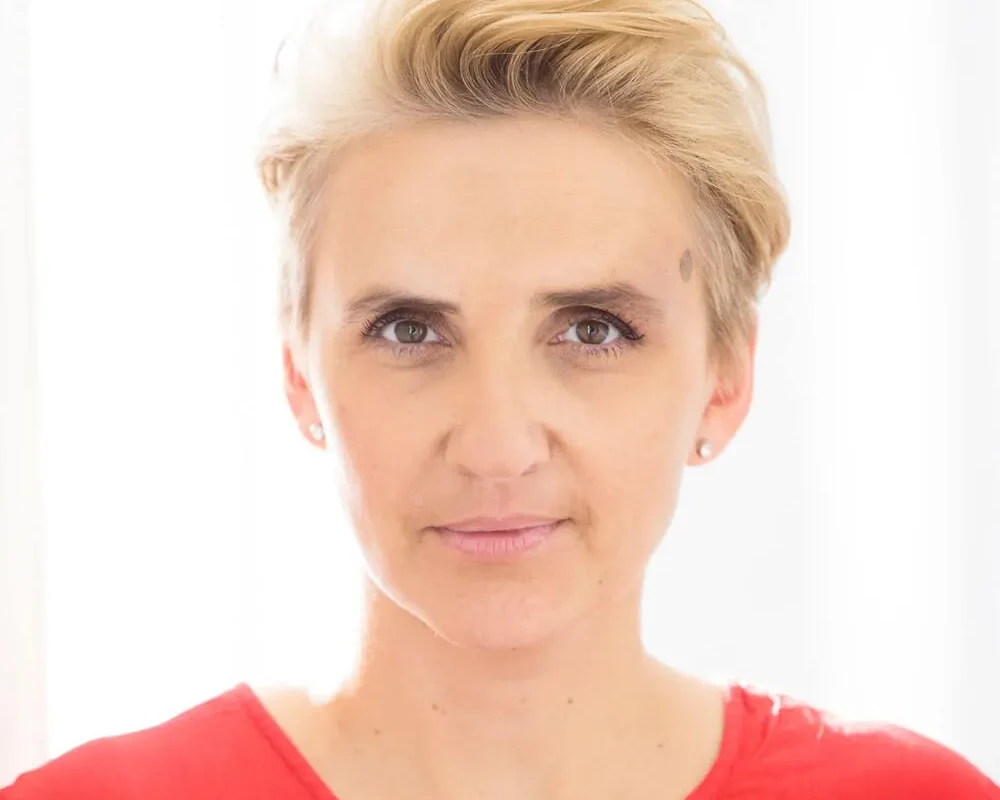 Kobieta w czerwonej bluzce z delikatnym uśmiechem patrzy prosto w obiektyw na tle jasnego okna.