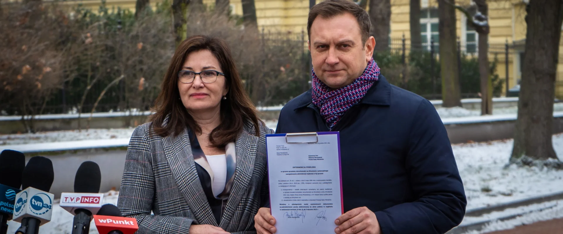Na zdjęciu widać dwie osoby stojące na zewnątrz, trzymające dokument, z mikrofonami mediów w tle, w zimowej scenerii.