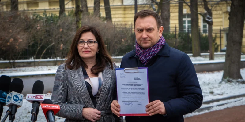 Na zdjęciu widać dwie osoby stojące na zewnątrz, trzymające dokument, z mikrofonami mediów w tle, w zimowej scenerii.