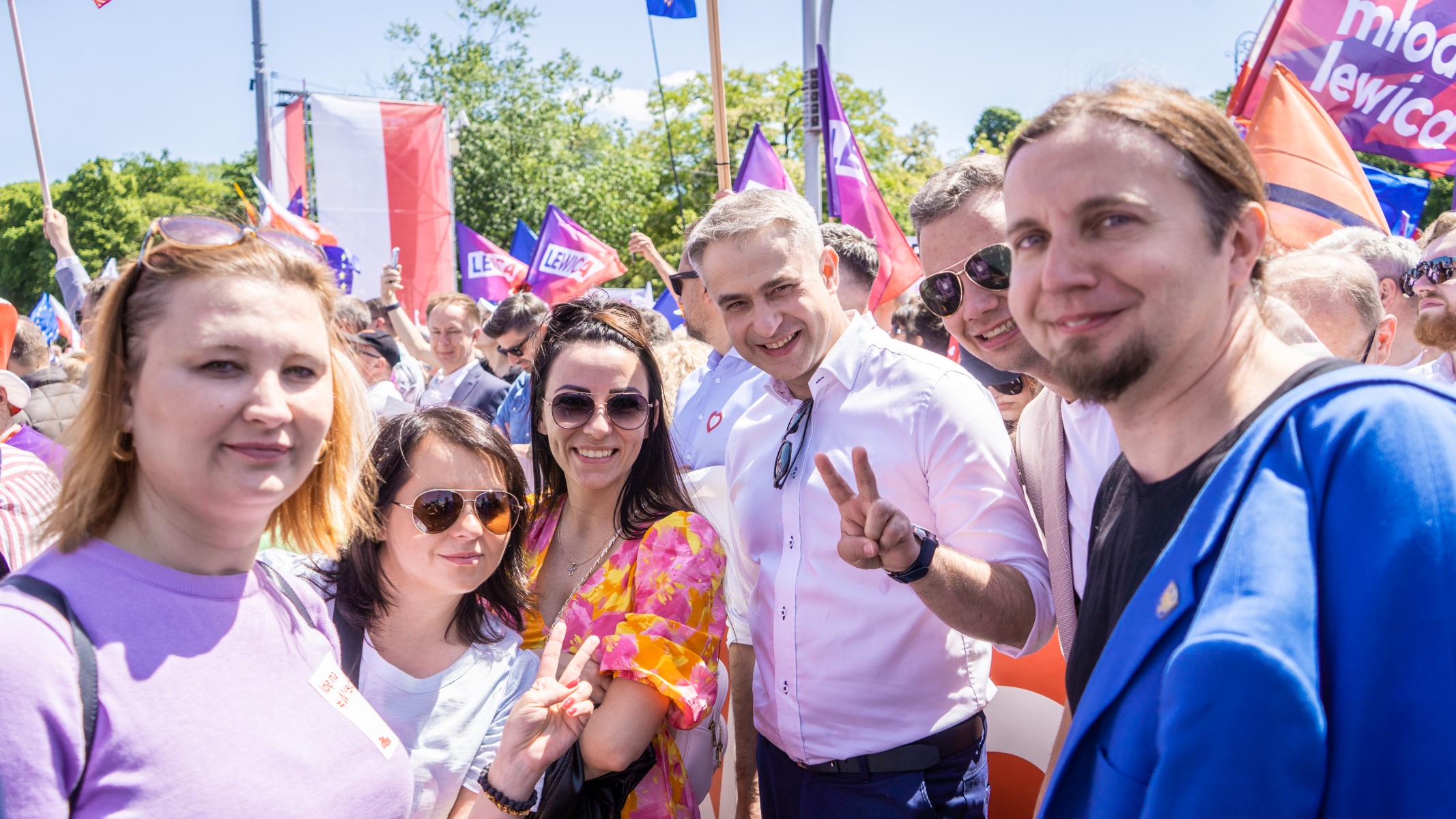 Grupa uczestników Marszu Lewicy 4 czerwca trzyma flagi i banery z napisem LEWICA, uśmiechnięta pozującą do zdjęcia w tłumie.