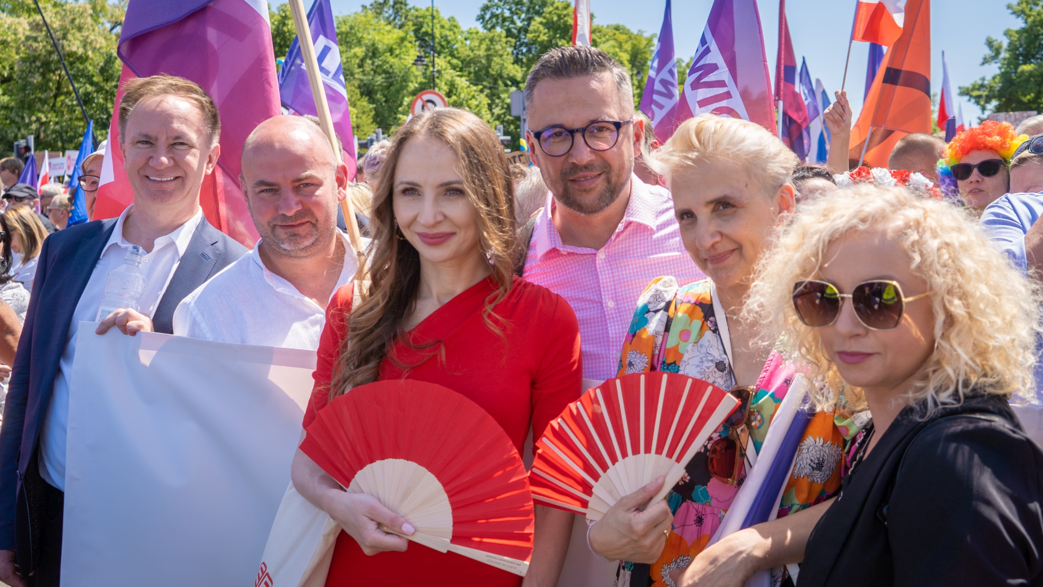Grupa uczestników Lewicy na Marszu 4 czerwca, stojąca razem i uśmiechnięta, trzyma czerwone wachlarze i flagi w różnych kolorach na tle tłumu w słoneczny dzień.