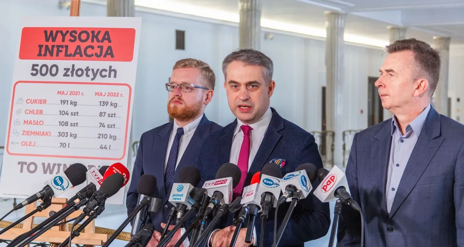 Trzech mężczyzn w garniturach podczas konferencji prasowej stoi obok planszy z napisem WYSOKA INFLACJA 500 złotych, pokazującej porównanie cen produktów spożywczych z 2021 i 2022.