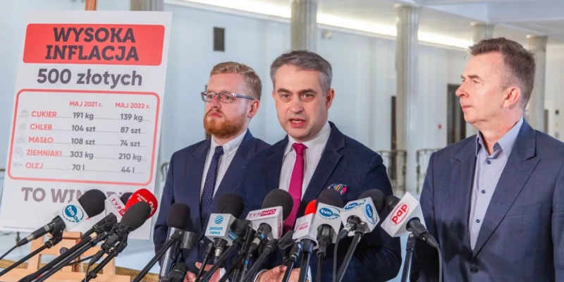 Trzech mężczyzn w garniturach podczas konferencji prasowej stoi obok planszy z napisem WYSOKA INFLACJA 500 złotych, pokazującej porównanie cen produktów spożywczych z 2021 i 2022.