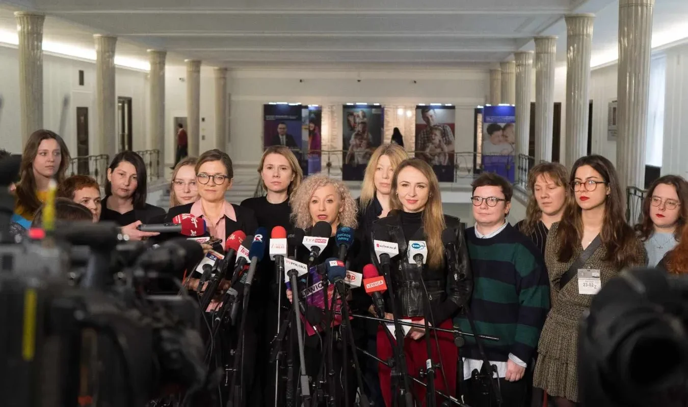 Grupa dziennikarek stoi w holu przed ustawionymi mikrofonami podczas konferencji prasowej.