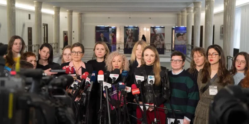 Grupa dziennikarek stoi w holu przed ustawionymi mikrofonami podczas konferencji prasowej.