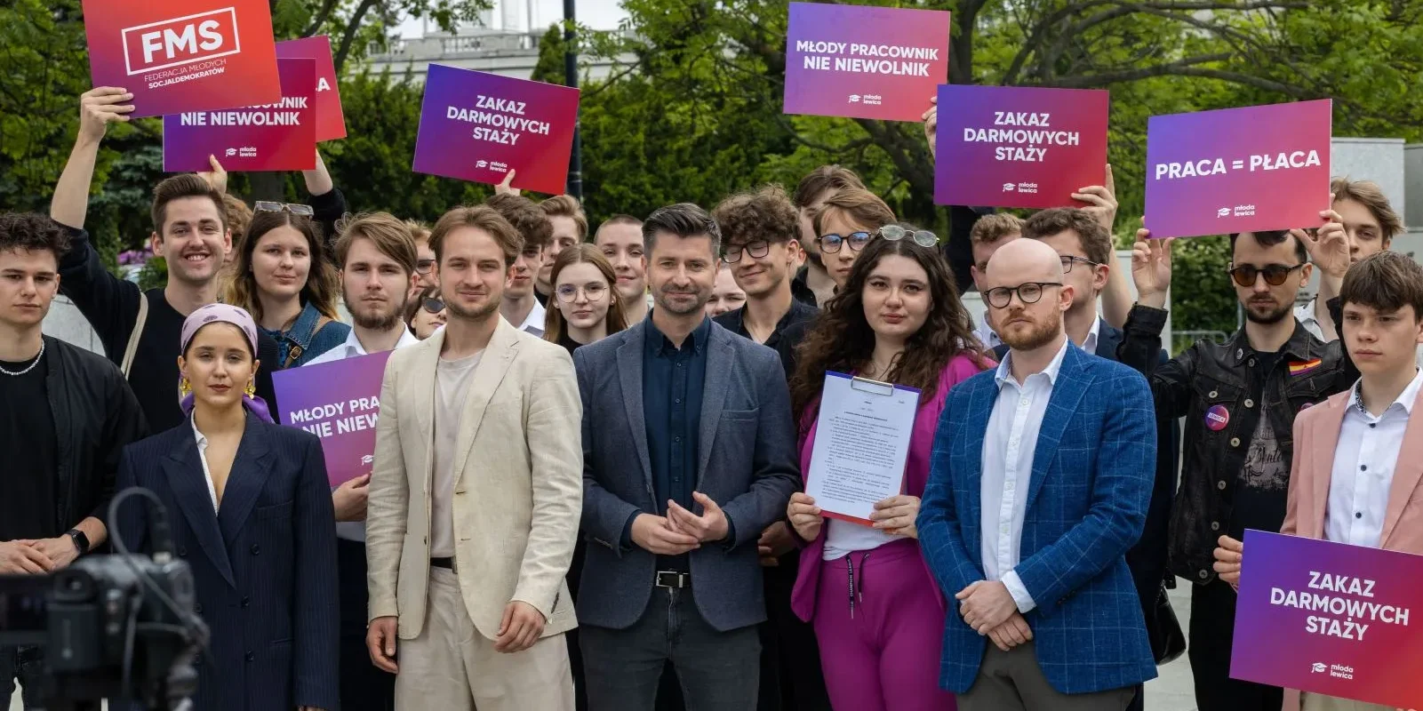 Grupa młodych ludzi bierze udział w demonstracji, trzymając kolorowe plakaty z hasłami „Zakaz darmowych staży” i „Praca = Płaca”.