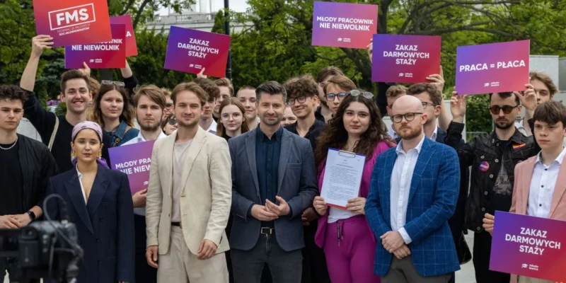 Grupa młodych ludzi bierze udział w demonstracji, trzymając kolorowe plakaty z hasłami „Zakaz darmowych staży” i „Praca = Płaca”.