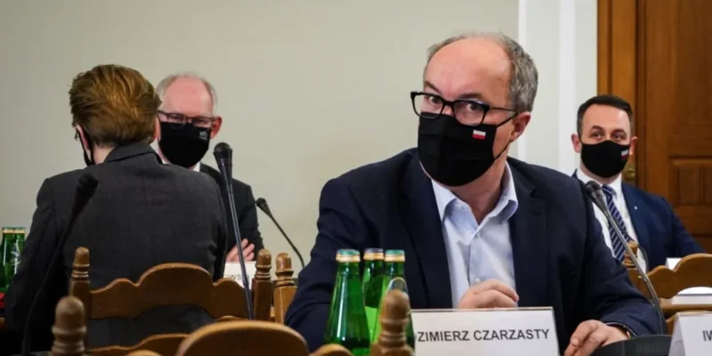 Grupa osób w garniturach podczas posiedzenia; na pierwszym planie mężczyzna w okularach i czarnej maseczce siedzi przy stole z mikrofonem, obok widoczne zielone butelki.