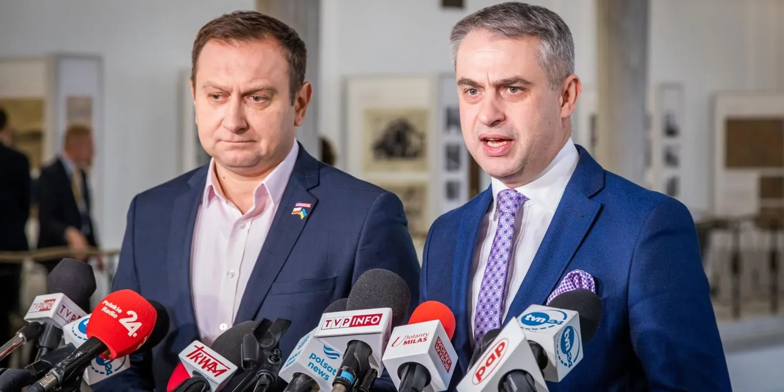 Dwóch mężczyzn w garniturach stoi przed mikrofonami, prowadząc konferencję prasową w jasnym wnętrzu.