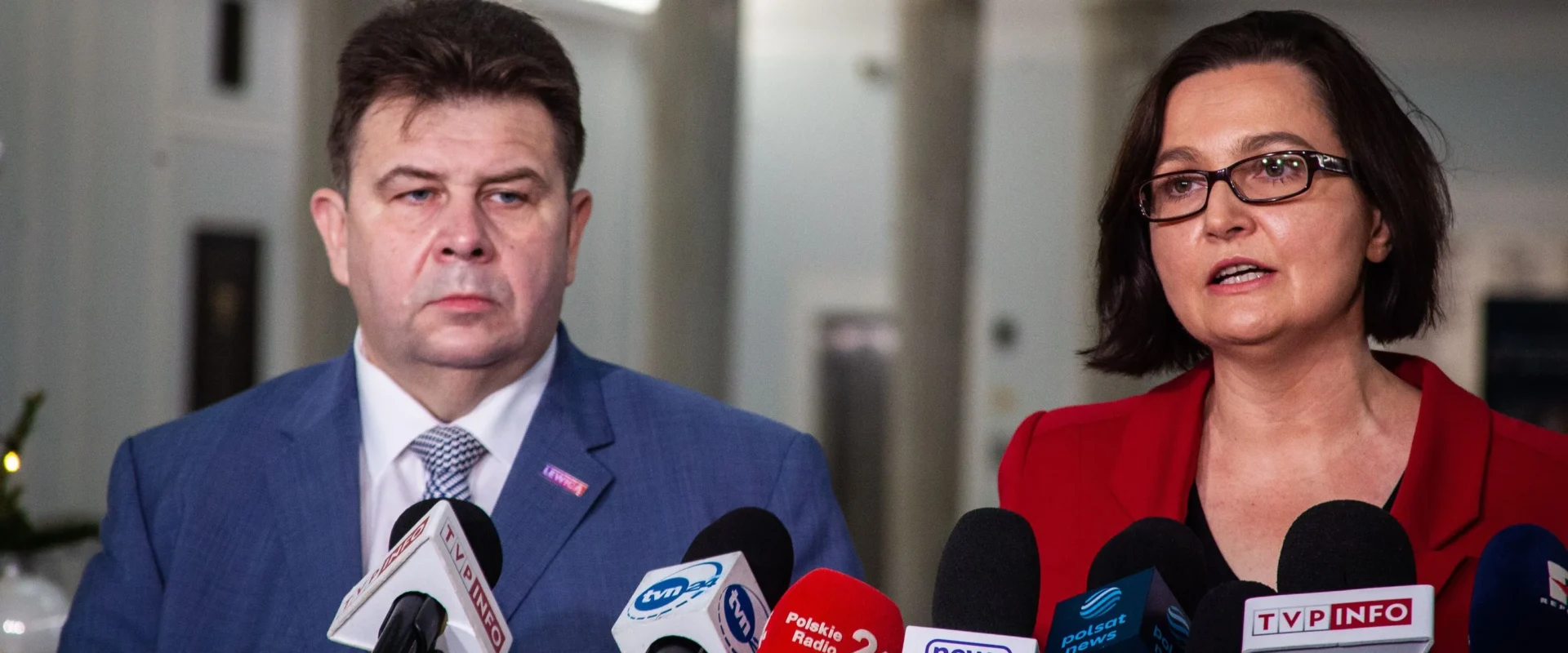 Dwóch polityków stoi przy mikrofonach podczas konferencji prasowej, otoczonych logotypami mediów.