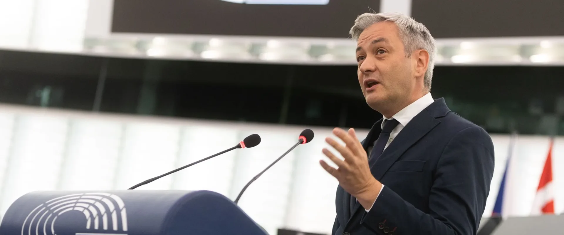 Mężczyzna w ciemnym garniturze przemawia z mikrofonami przy mównicy Parlamentu Europejskiego, na pulpicie widnieje logo UE, a w tle widać flagi państw członkowskich.