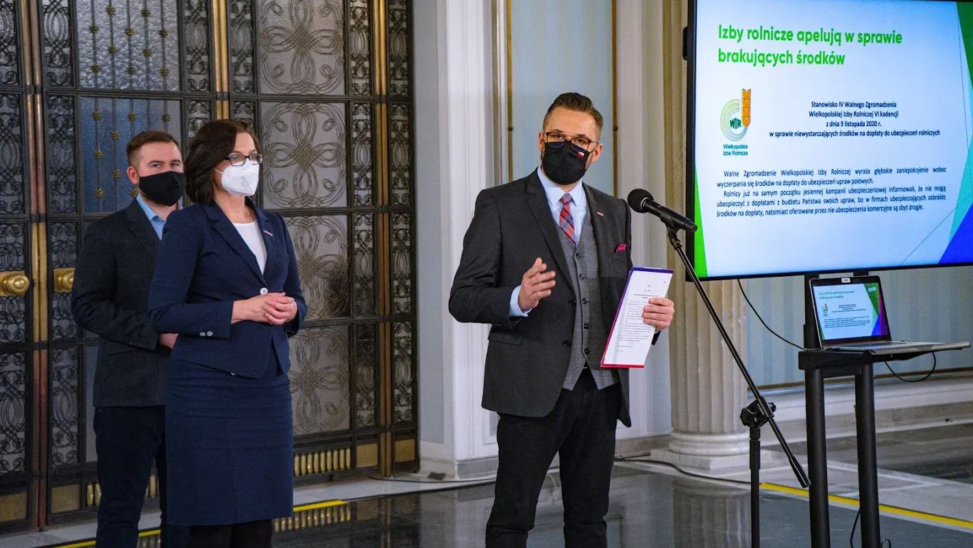 Trzy osoby w maseczkach podczas konferencji prasowej, prowadzący z mikrofonem i notatnikiem, w tle slajd dotyczący dopłat do ubezpieczeń upraw i hodowli.