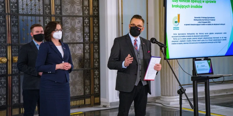 Trzy osoby w maseczkach podczas konferencji prasowej, prowadzący z mikrofonem i notatnikiem, w tle slajd dotyczący dopłat do ubezpieczeń upraw i hodowli.