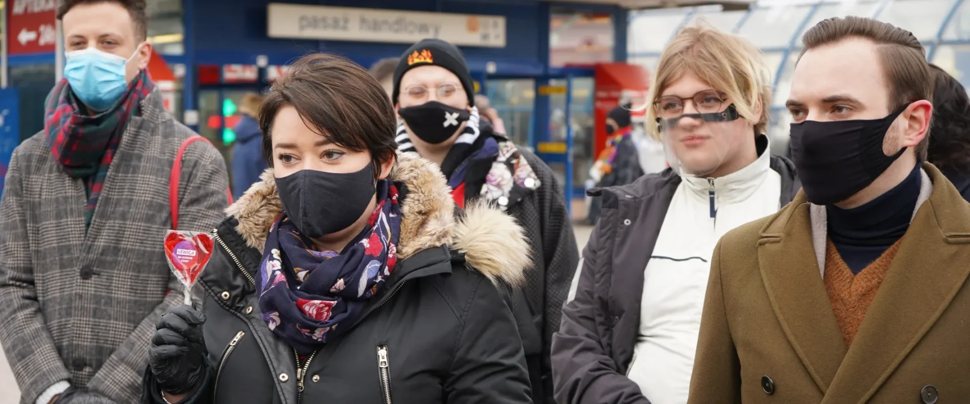 Grupa osób w maseczkach na zewnątrz sklepu; na pierwszym planie kobieta w czarnej kurtce z futrzanym kołnierzem trzyma lizak w kształcie serca.