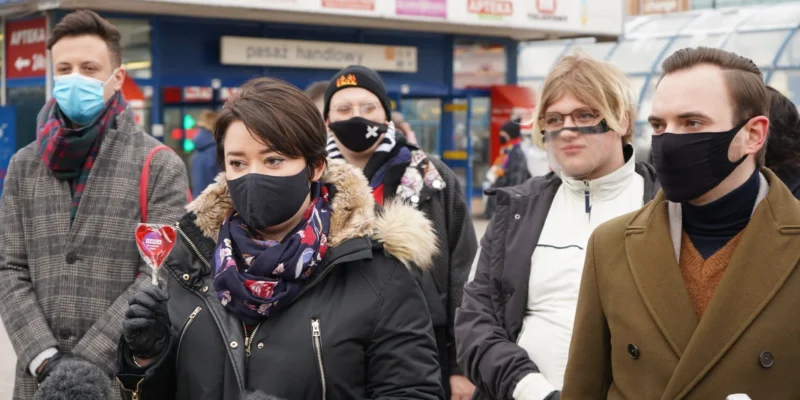 Grupa osób w maseczkach na zewnątrz sklepu; na pierwszym planie kobieta w czarnej kurtce z futrzanym kołnierzem trzyma lizak w kształcie serca.