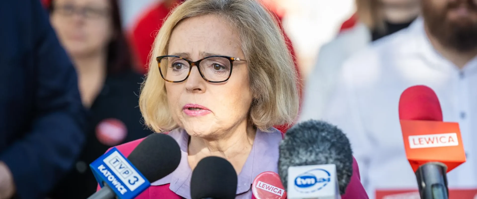 Kobieta w okularach, ubrana w różowy żakiet, przemawia do mediów podczas konferencji prasowej, otoczona mikrofonami z logo Lewicy.