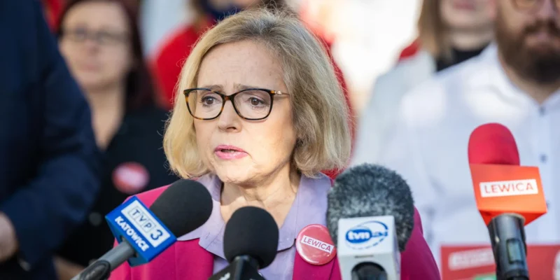 Kobieta w okularach, ubrana w różowy żakiet, przemawia do mediów podczas konferencji prasowej, otoczona mikrofonami z logo Lewicy.