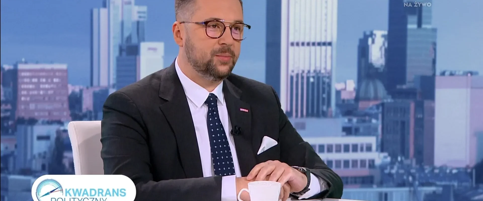 Mężczyzna w garniturze i okularach siedzi w studiu telewizyjnym, na stole ma filiżankę kawy i telefon, w tle widać panoramę miasta oraz logo programu „Kwadrans Polityczny”.
